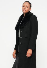 Cappotto nero con colletto in pelliccia sintetica, caratterizzato da un design lungo, cinque bottoni e tasche frontali. Il materiale ha una texture liscia.