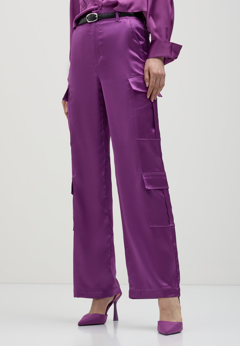 Lila Satin- Cargo-Hosen mit seitlichen Taschen, schmaler Schnitt und weitem Bein. Getragen mit einem schwarzen Gürtel und passenden lila High Heels. Glatte Textur.