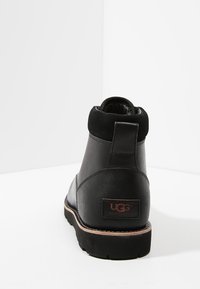 Botas de cuero negras hasta el tobillo con un acabado texturizado, punta redondeada y un tacón bajo; presentan un parche de marca en la parte posterior.