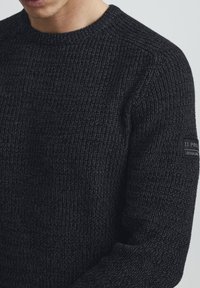 Maglione nero lavorato a maglia con texture a coste, dotato di scollatura a girocollo e di un'etichetta con marchio sulla manica sinistra.