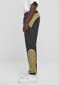 Træningsbukser i sort stof med et kontrastpanel i khaki, hvid kant og elastisk talje. Båret med hvide sneakers.