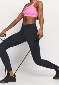 Różowy stanik sportowy Nike, czarne legginsy z wysokim stanem i czarne buty sportowe. Osoba trzyma gumy oporowe, prezentując design odzieży sportowej.