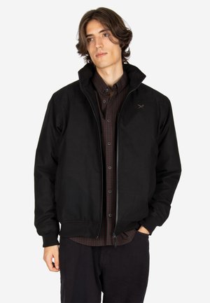 Junger Mann mit mittelbraunem Haar, der eine schwarze Reißverschlussjacke über einem dunklen karierten Hemd trägt und mit einer Hand in der Tasche steht.