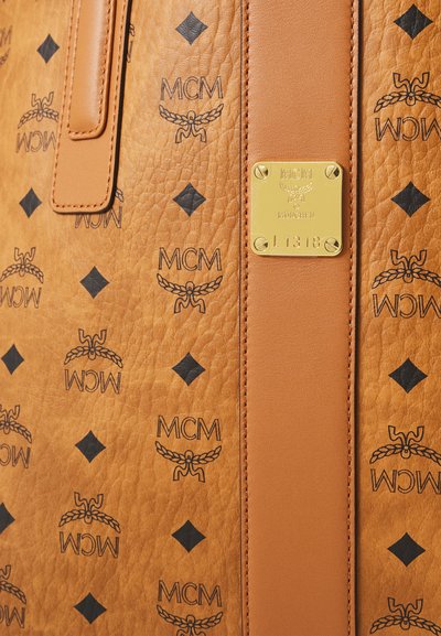 Tan leather textured backpack with repeated black MCM logo and diamond patterns, featuring a vertical leather strap and gold brand plaque.
