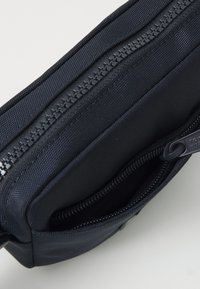 Czarna nylonowa saszetka z podwójnym zapięciem na zamek. Teksturowana powierzchnia, kompaktowy design oraz dodatkowa kieszeń wewnętrzna. Trwała i lekka.