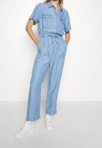 Person som bär en ljusblå denimoverall med korta ärmar, framfickor och vita sneakers, ståendes med händerna i fickorna.