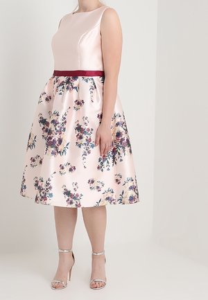 Femme portant une robe sans manches en satin rose pâle avec une ceinture bordeaux à la taille et une jupe fleurie, chaussée de talons argentés à bride à la cheville.