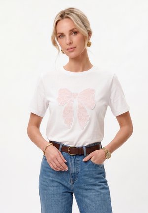 Kvinde iført hvid t-shirt med pink blonder sløjfe design, blå jeans, brun bælte, guldur, armbånd og runde guldøreringe, stående mod hvid baggrund.