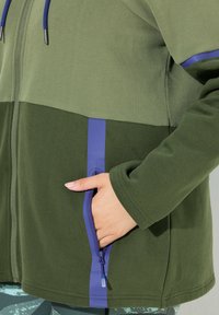 Green zip-up jacket with a color-block design, featuring a lighter green upper section and dark green lower section. Blue zipper accents.