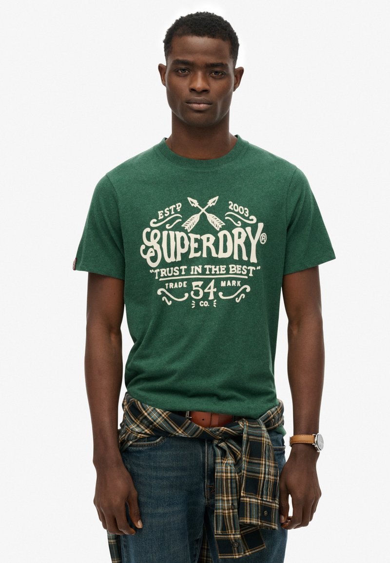 Grøn bomulds t-shirt med hvid Superdry-logo og tekst, med rund halsudskæring og korte ærmer. Bæres med blå jeans og en ternet skjorte knyttet omkring taljen.