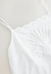 Caraco blanc avec fines bretelles doubles, bordure en dentelle festonnée le long de l'encolure et broderie florale en dentelle complexe sur le devant.