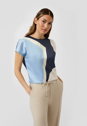 Frau trägt ein kurzärmeliges Oberteil mit blauen, marineblauen und cremefarbenen geometrischen Formen, kombiniert mit beigen Hosenzügen, Hände in den Taschen.