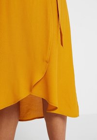 Abito avvolgente giallo realizzato in tessuto testurizzato, con un design fluido, un orlo leggermente asimmetrico e dettaglio con lacci sul lato.