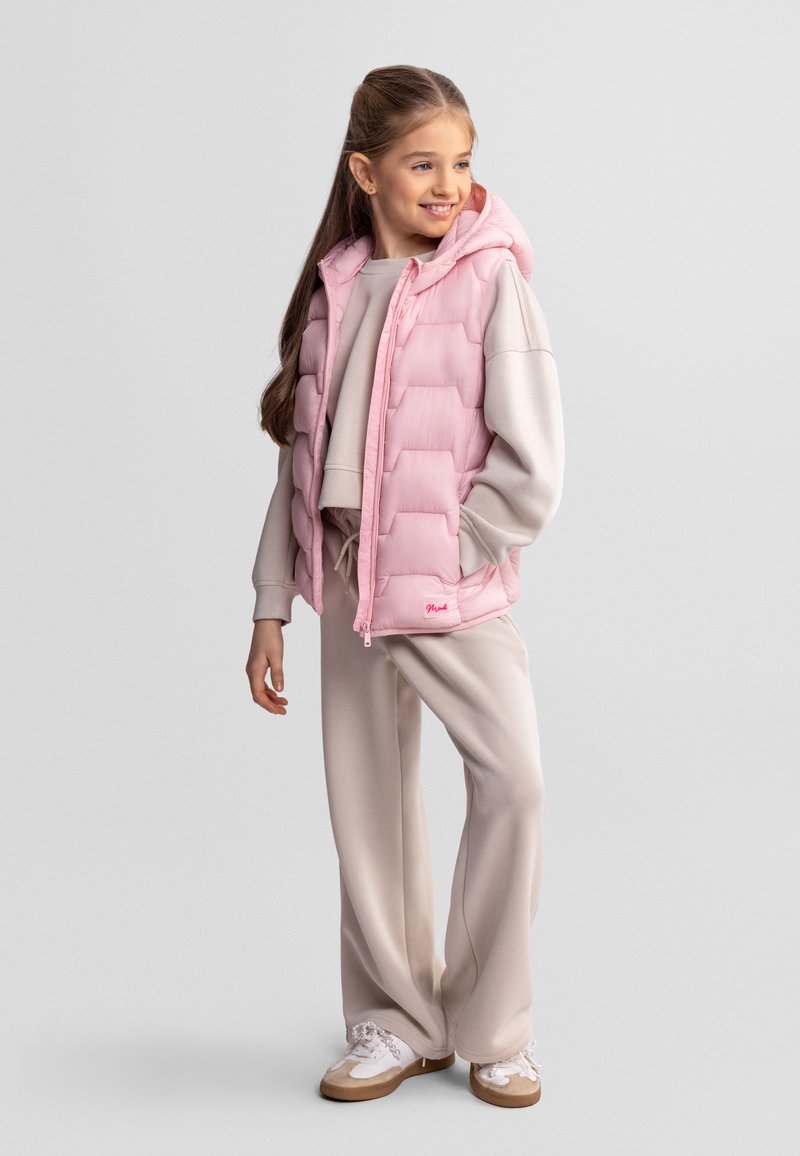 Jeune fille souriante, portant une doudoune sans manches rose clair sur un sweat-shirt et un pantalon beiges, debout avec une main dans la poche et des baskets aux pieds.