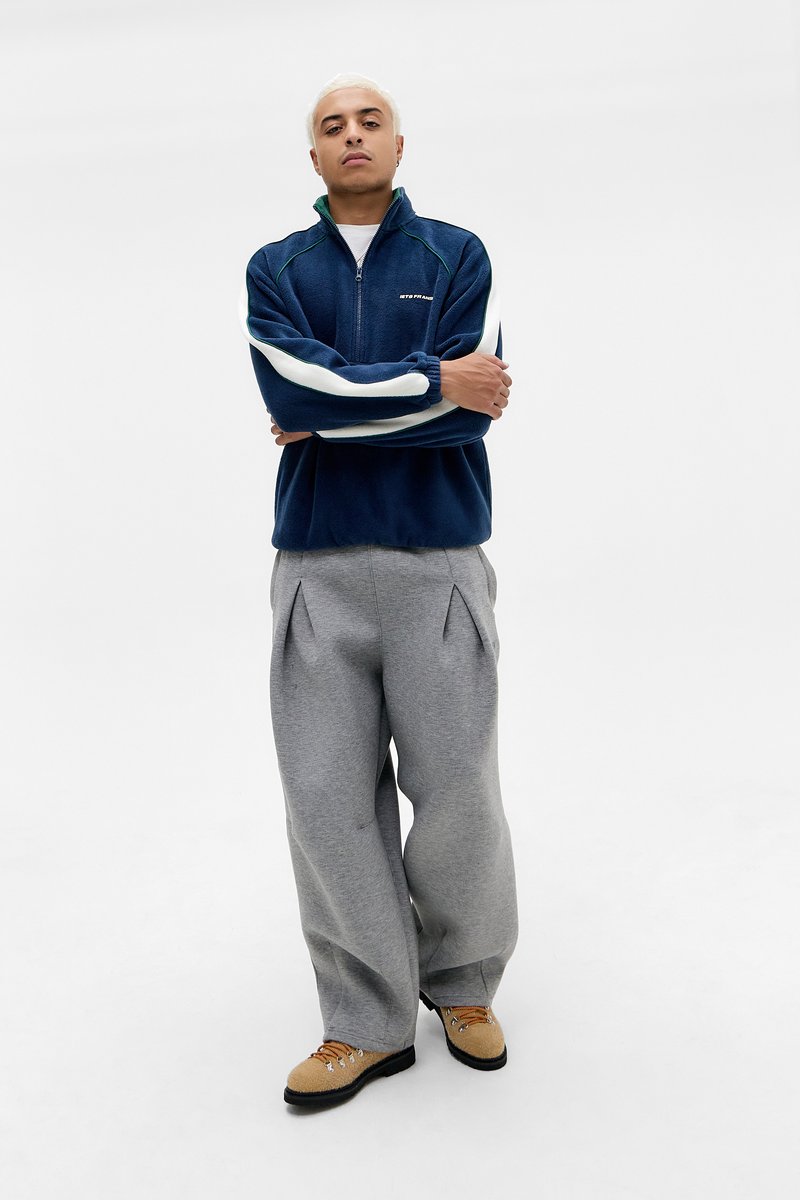 Giacca a zip in pile blu navy con accenti verdi e bianchi sulle maniche, abbinata a pantaloni della tuta grigi a gamba larga e scarpe ispirate all'escursionismo color beige.