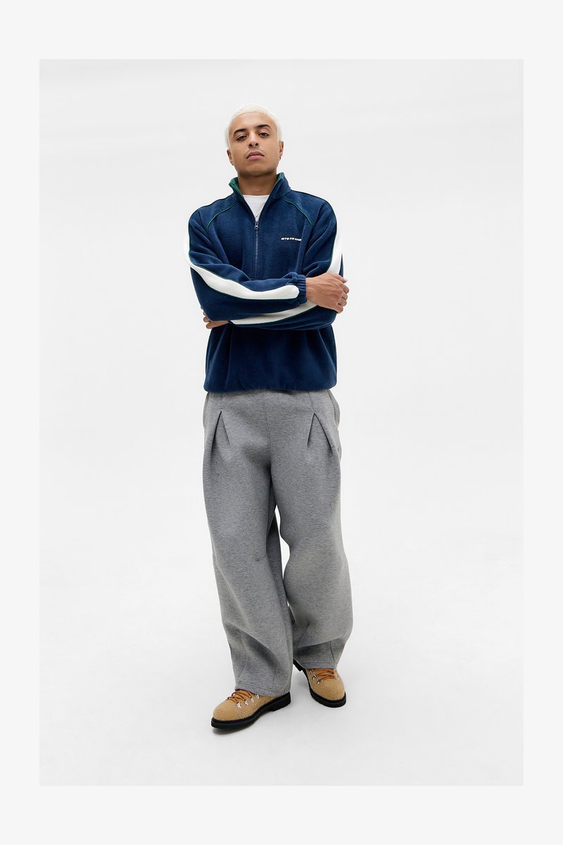 Giacca a zip in pile blu navy con accenti verdi e bianchi sulle maniche, abbinata a pantaloni della tuta grigi a gamba larga e scarpe ispirate all'escursionismo color beige.