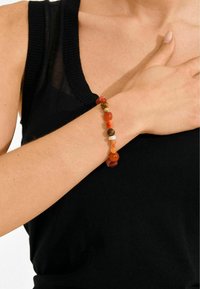 Pulsera de cuentas con una mezcla de gemas en tonos naranja, marrón y crema. Se lleva en la muñeca, combinada con una camiseta negra sin mangas.