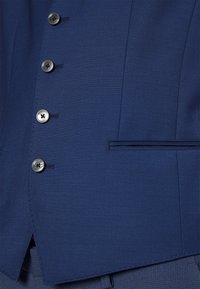 Vesta bleumarin cu un material texturat, cinci nasturi negri și o singură pungă cu pipă. Linii curate și detalii de design croite.
