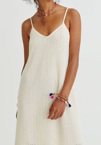 Vestido de ganchillo color crema con tirantes finos y escote en V, que presenta un patrón texturizado. Pulsera multicolor con borlas en la muñeca.