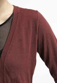 Burgunderfarbener Cardigan mit V-Ausschnitt und gerippter Textur, über einem schwarzen Mesh-Top getragen, das die kontrastierenden Stoffe und Stile hervorhebt.