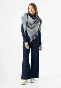 Grau gemischter Wollschal mit fransigen Rändern, über einem marineblauen Oberteil drapiert. Kombiniert mit weit geschnittenen, dunklen Jeans und hellen Sneakers.