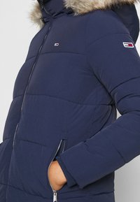 Marinblå vadderad jacka med huva med konstpälsdetalj, fickor med dragkedja och logotypdetalj p å ärmen. Slät yta, figursydd design.