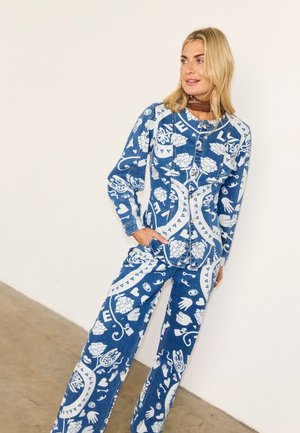 Γυναίκα που φοράει πουκάμισο με κουμπιά με μπλε και λευκό μοτίβο και αντίστοιχο παντελόνι φαρδιάς γραμμής, στέκεται μπροστά σε έναν λευκό τοίχο με τα χέρια στις τσέπες.