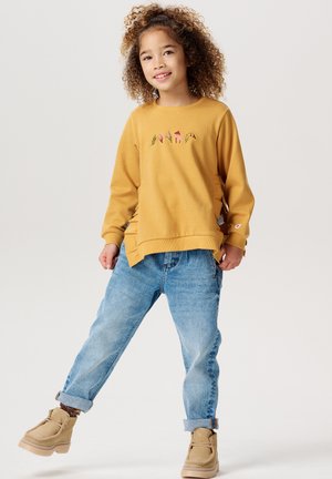 Senfgelbes Sweatshirt mit gestickten bunten Mustern, kombiniert mit hellblauen, umgeschlagenen Jeans und braunen Stiefeln. Weicher Stoff, lässige Passform.