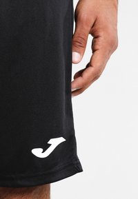 Czarne sportowe spodenki z gładkiego materiału, z białym logo przy krawędzi, dopasowany krój i czyste przeszycia.