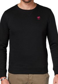 Czarny sweatshirt z okrągłym dekoltem, o fakturze ribbed i dopasowanym kroju. Posiada małe różowe logo na piersi oraz ściągacze na rękawach i u dołu.