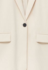 Blazer crema con un solo botón negro, solapas con muesca y dos bolsillos frontales con una textura suave. El diseño entallado ofrece un ajuste estructurado.