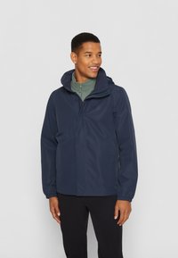 Veste imperméable marine avec une capuche ajustable, fermeture à glissière sur le devant et poignets élastiques. Portée par-dessus une couche intérieure zippée de couleur vert clair.