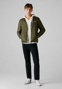 Chaqueta de plumas verde oliva con cierre de cremallera, tejido texturizado y una sudadera blanca debajo. Combinada con jeans oscuros y zapatillas blancas.