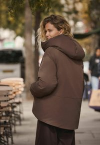 Bruine gewatteerde jas met een grote capuchon, met een soepele textuur en subtiele glans. Van achteren gezien, gecombineerd met donkere broeken.