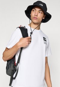 White T-shirt with logo, worn under a black bucket hat. User carries a dark gray backpack with red accents and adjustable straps.