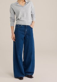 Harmaa neulepaita V-kaula-aukolla, yhdistettynä leveisiin sinisiin farkkuihin. Farkuissa on klassinen leikkaus ja hieman kulumista. Ruskeat kengät.