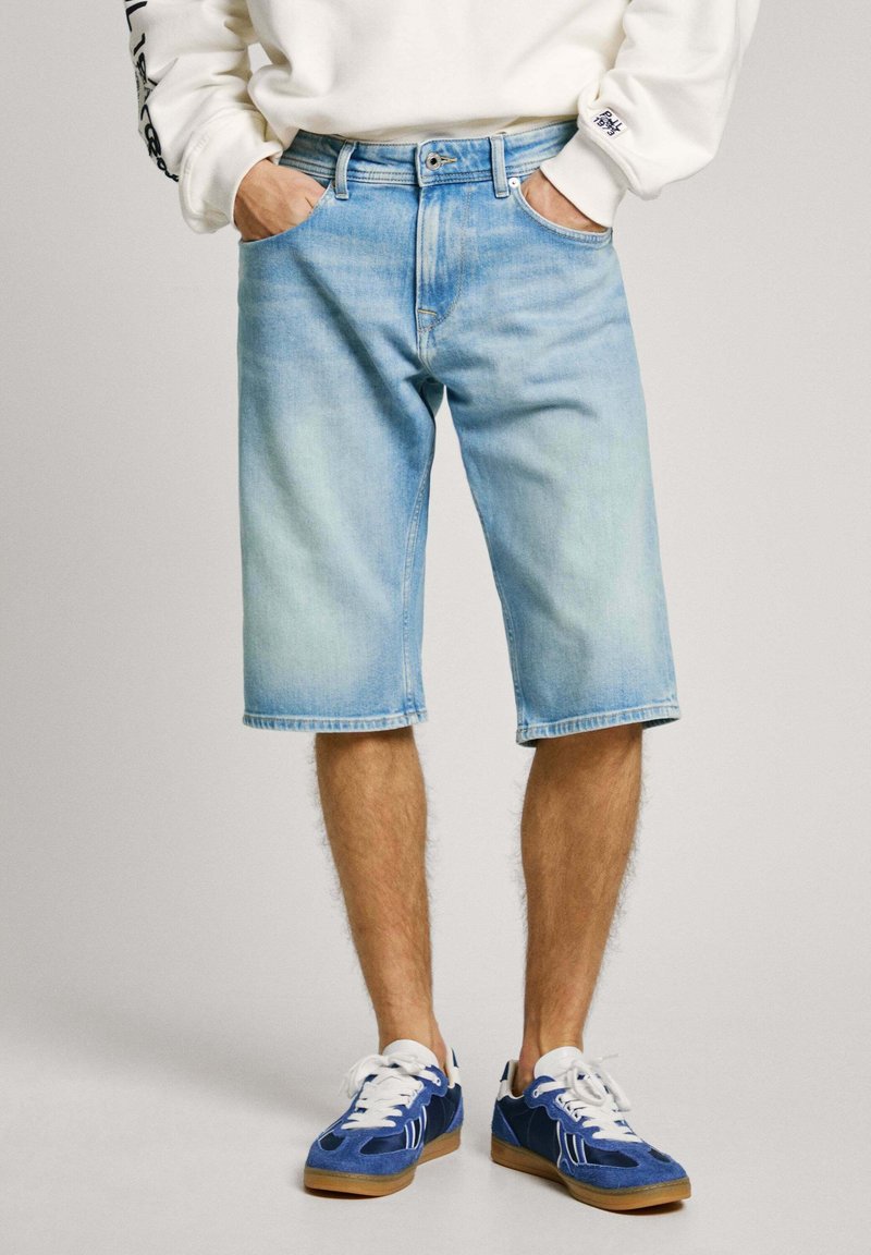 Pantalones cortos de mezclilla azul claro con un corte relajado, lazos para cinturón y bolsillos delanteros; combinados con zapatillas azules y una camiseta blanca de manga larga.