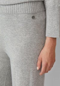 Gros plan sur une personne portant un pantalon en tricot gris clair et un pull, avec un petit logo métallique rond visible près de la taille et une main détendue.