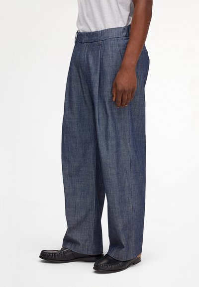 Man wearing loose-fitting blue denim trousers, black loafers, and a light gray tucked-in shirt, standing against white background.