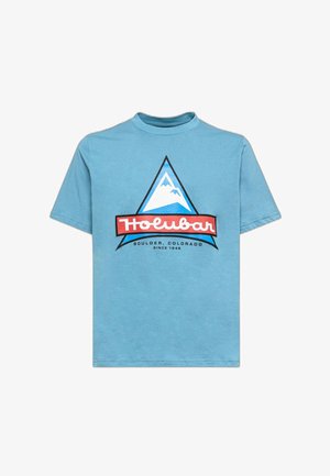 T-shirt en coton bleu clair avec un col rond. Présente un graphique triangulaire de montagne en blanc et bleu, ainsi qu'un logo rouge "Holubar" à la base.