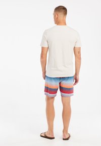 Lichte beige t-shirt voor mannen, gecombineerd met multicolore zwemshorts met horizontale strepen in blauw, oranje en rood. Bruine sandalen.