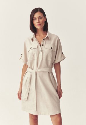 Kobieta w beżowej sukience koszulowej z paskiem, krótkimi rękawami i zapinanymi na guziki kieszeniami na piersiach, stojąca na tle jednolitego jasnego tła.