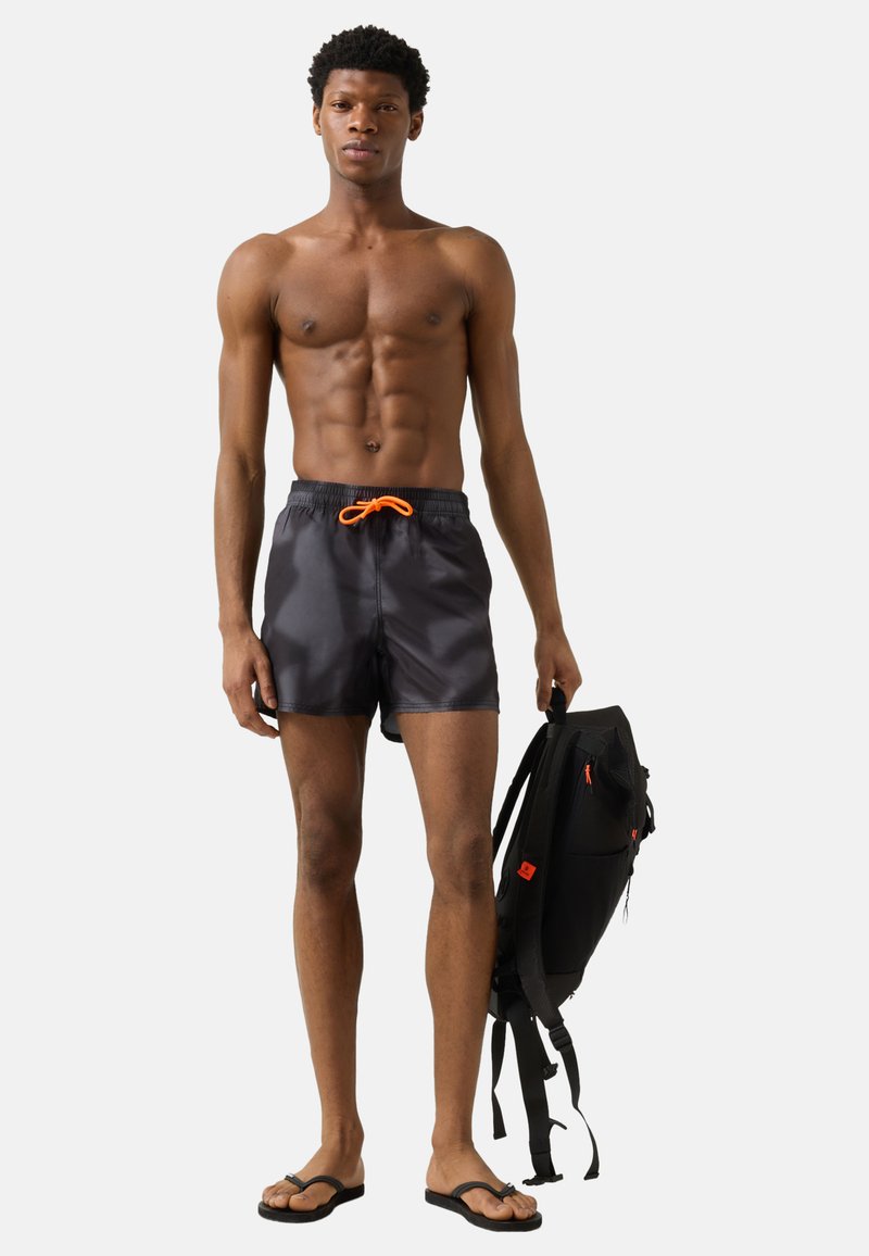 Homme aux muscles toniques portant un short de bain noir avec un cordon orange, des tongs noires, tenant un sac à dos noir avec des accents orange.