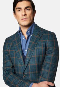 Zielono-niebieska marynarka w kratkę z wełny tweedowej, z klapami, w połączeniu z jasnoniebieską koszulą z guzikiem. Posiada dwa guziki oraz teksturowany materiał.