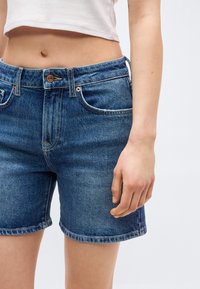 Γυναίκα που φοράει μπλε τζιν σορτς μεσαίου ύψους και λευκό κοντό τοπ, σε απλό φωτεινό φόντο.