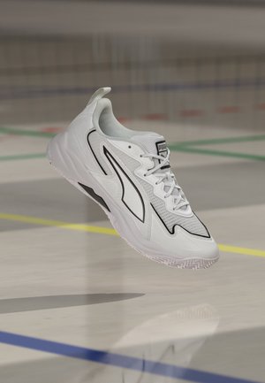 Hvid Puma sportssko med sorte accenter hængende over et indendørs banegulv markeret med farvede linjer.