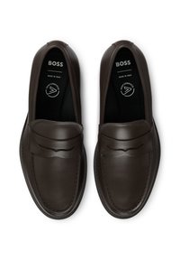 Zapatos de cuero de color marrón oscuro, sin cordones, con un acabado suave, que cuentan con una correa decorativa y costuras en contraste. Hechos en Italia.