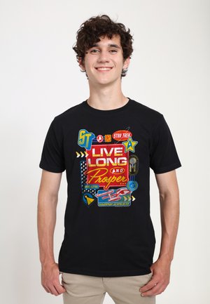 Jeune homme souriant portant un T-shirt noir avec un motif coloré "Star Trek Live Long and Prosper" et des icônes, pantalon beige, fond uni.