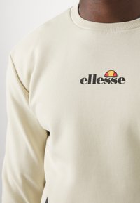 Světle béžová mikina vyrobená z měkké tkaniny, s kulatým výstřihem, pružnými manžetami a černým logem "ellesse" s barevným emblémem.