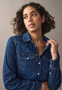Cămașă din denim albastru închis, cu guler clasic, două buzunare pe piept, nasturi cu capsenă și detalii de cusătură vizibile pe mâneci și corp.